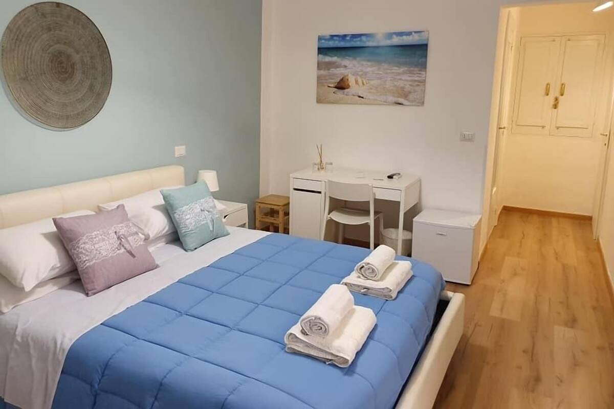 Un dormitori amb un llit, un escriptori i una pintura d'una escena de platja.