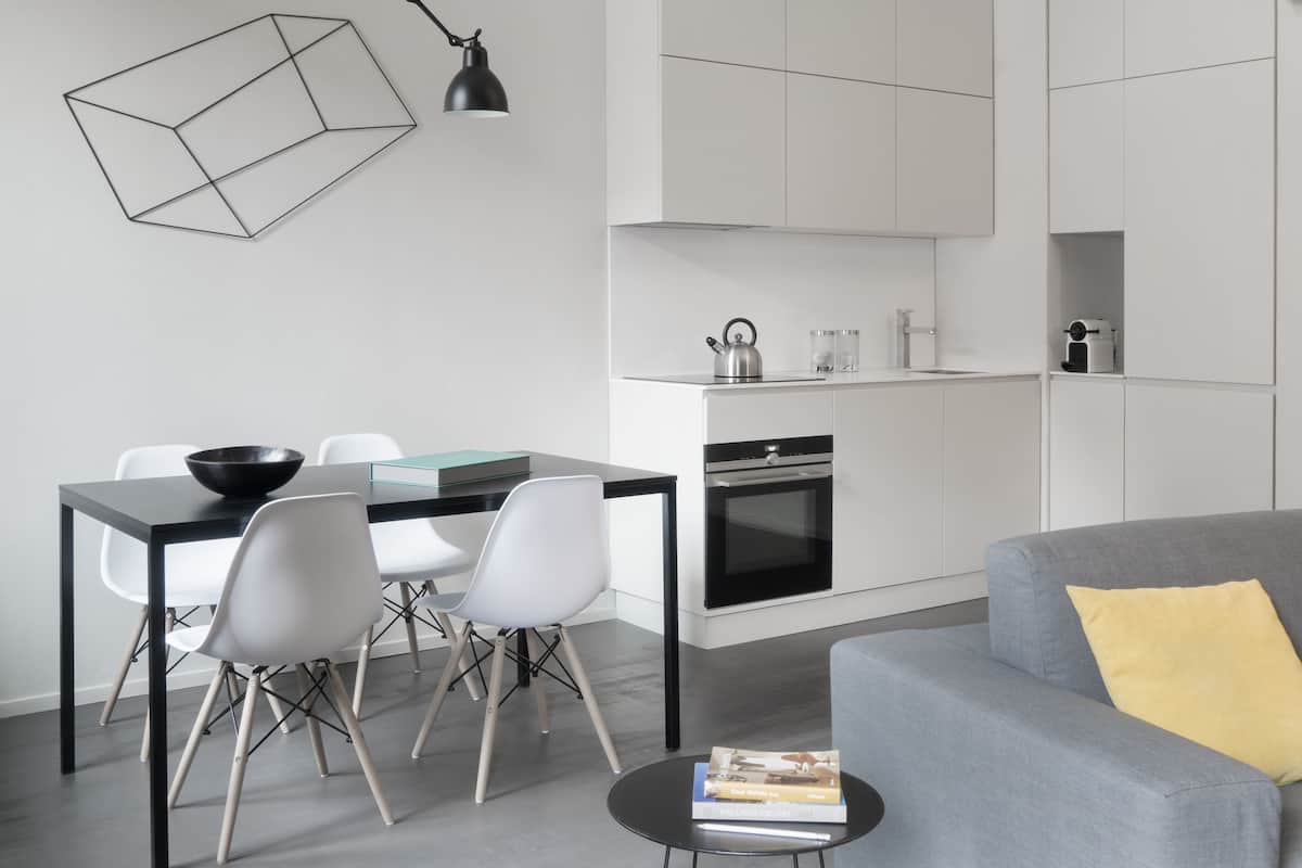 A modern kitchen with white cabinets, a black countertop, and a kettle. There is a dining area with a black table and white chairs. A gray sofa with a yellow pillow is in the foreground.
