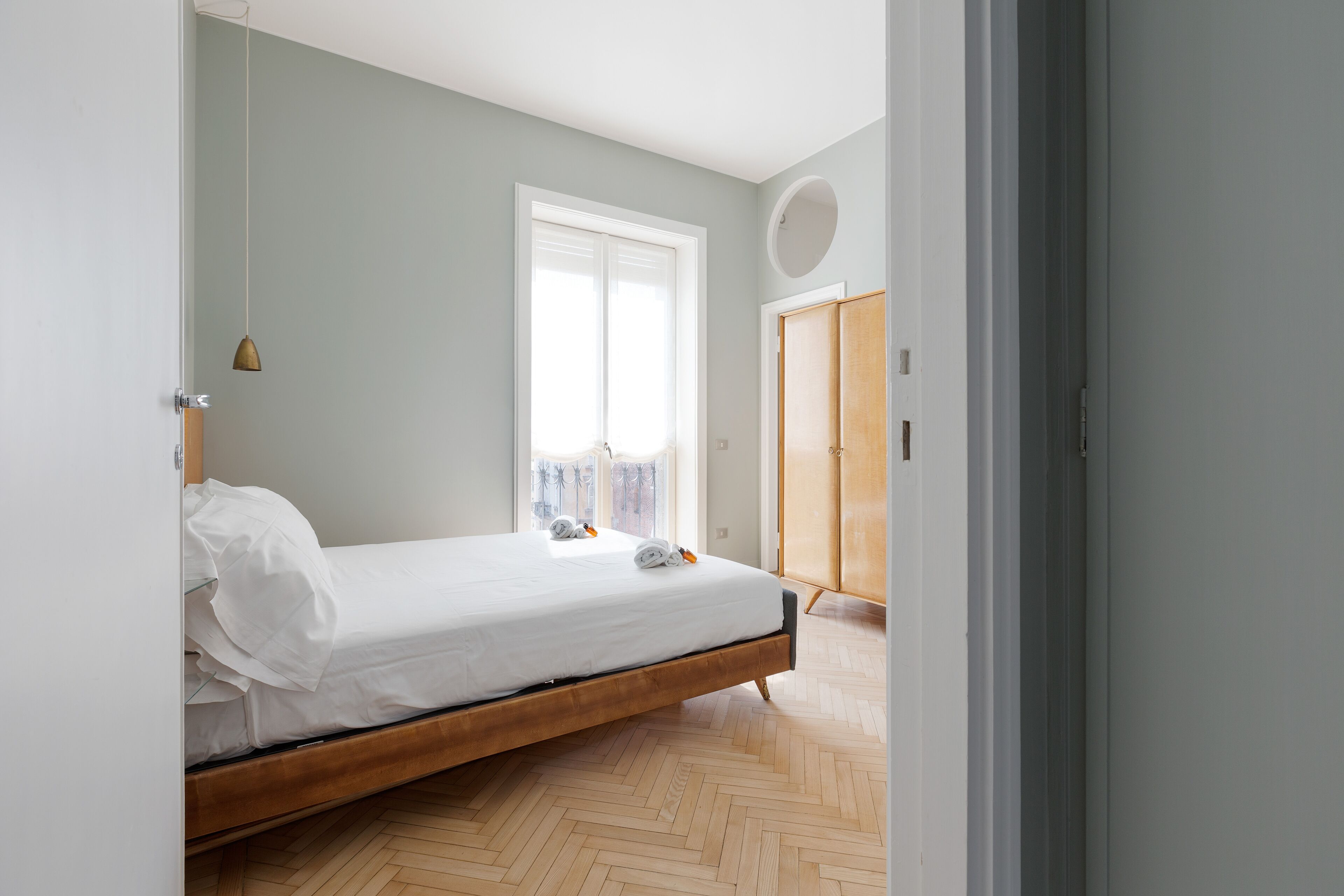 A minimalist bedroom with a wooden bed frame, a large window with curtains, and a wardrobe.