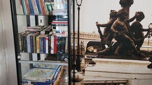 Uma vitrine de vidro com livros e jogos de quebra-cabeça, uma lousa com a palavra 'welcome' escrita e um abajur decorativo.