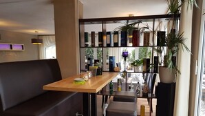 Um interior moderno com uma mesa de madeira, banquetas de bar, um sofá preto e uma estante repleta de livros e objetos decorativos.