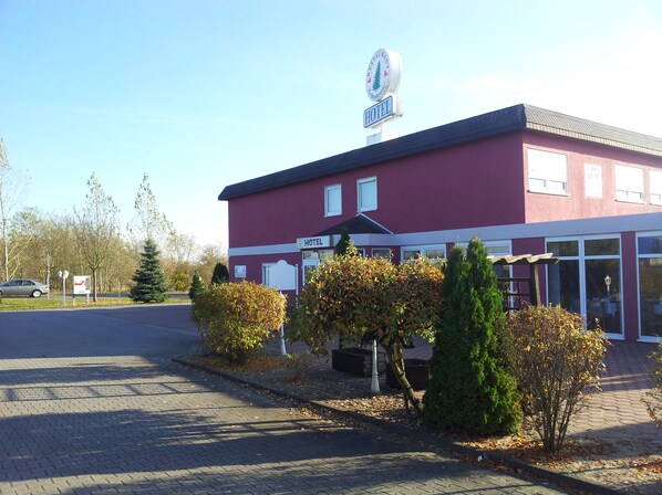 A red building with a sign indicating it is a hotel. There are trees and shrubs in front of the building. - Hotel zur Fichtenbreite Garni (Coswig)
