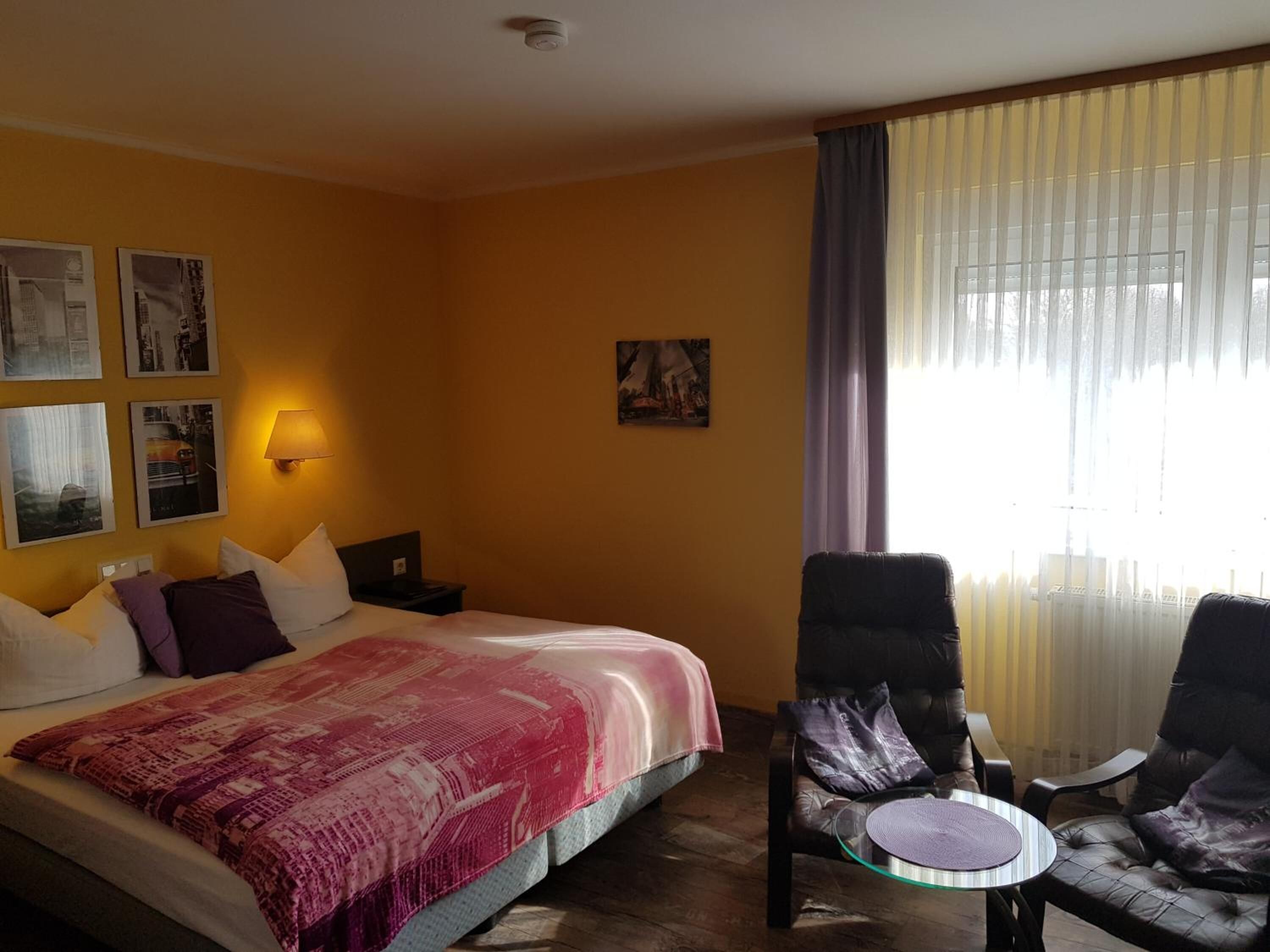 Ein Hotelzimmer mit einem Bett, einem Nachttisch, einem Sessel, einem Tisch und einem Fenster mit Vorhängen.