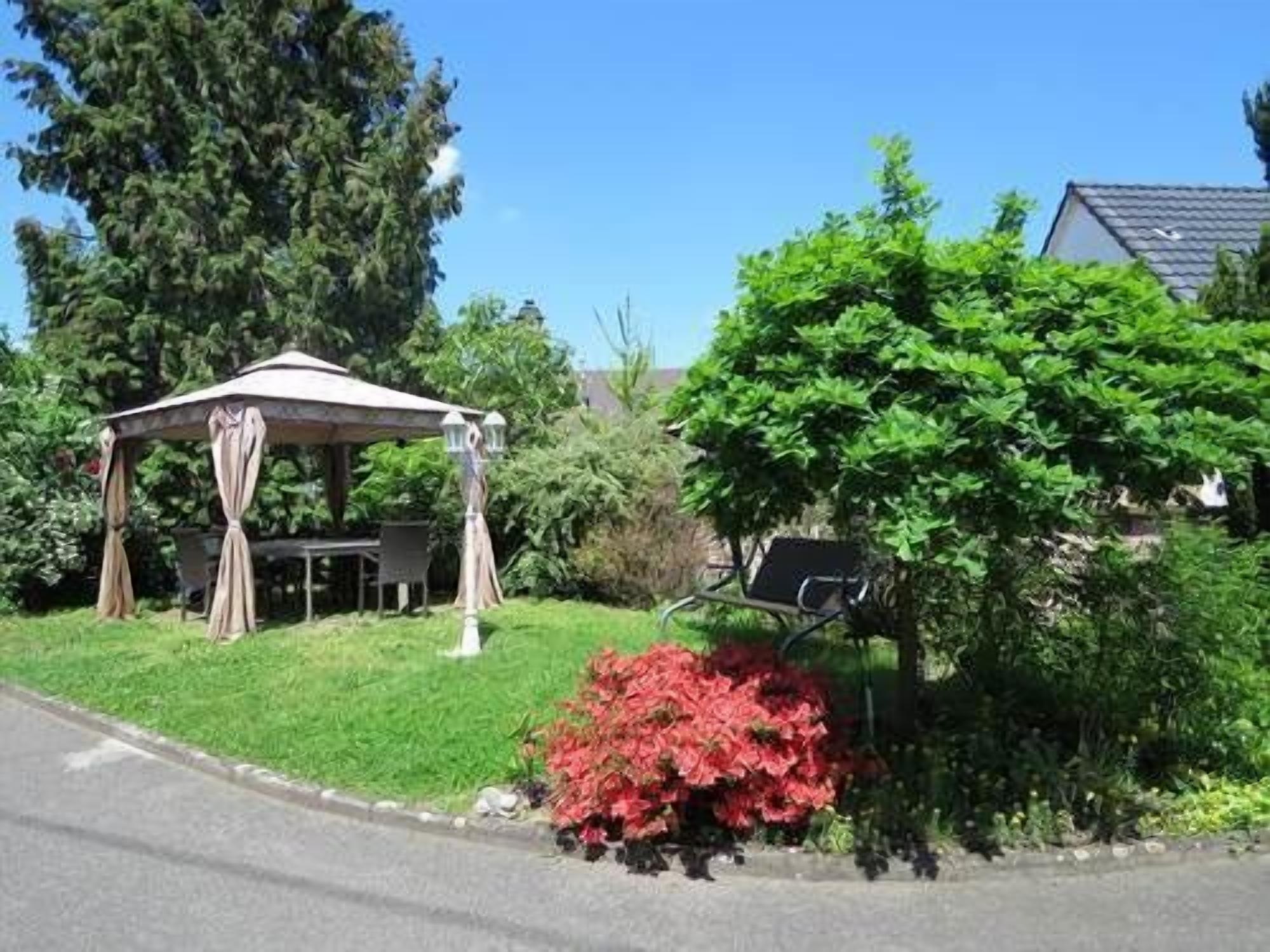 A gazebo with a table and chairs, surrounded by greenery and a red flowering bush.