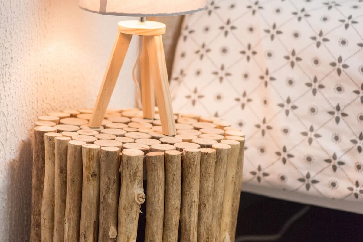 Ein Holznachttisch mit einer Lampe darauf, platziert vor einer Wand mit einem gemusterten Kissen im Hintergrund.