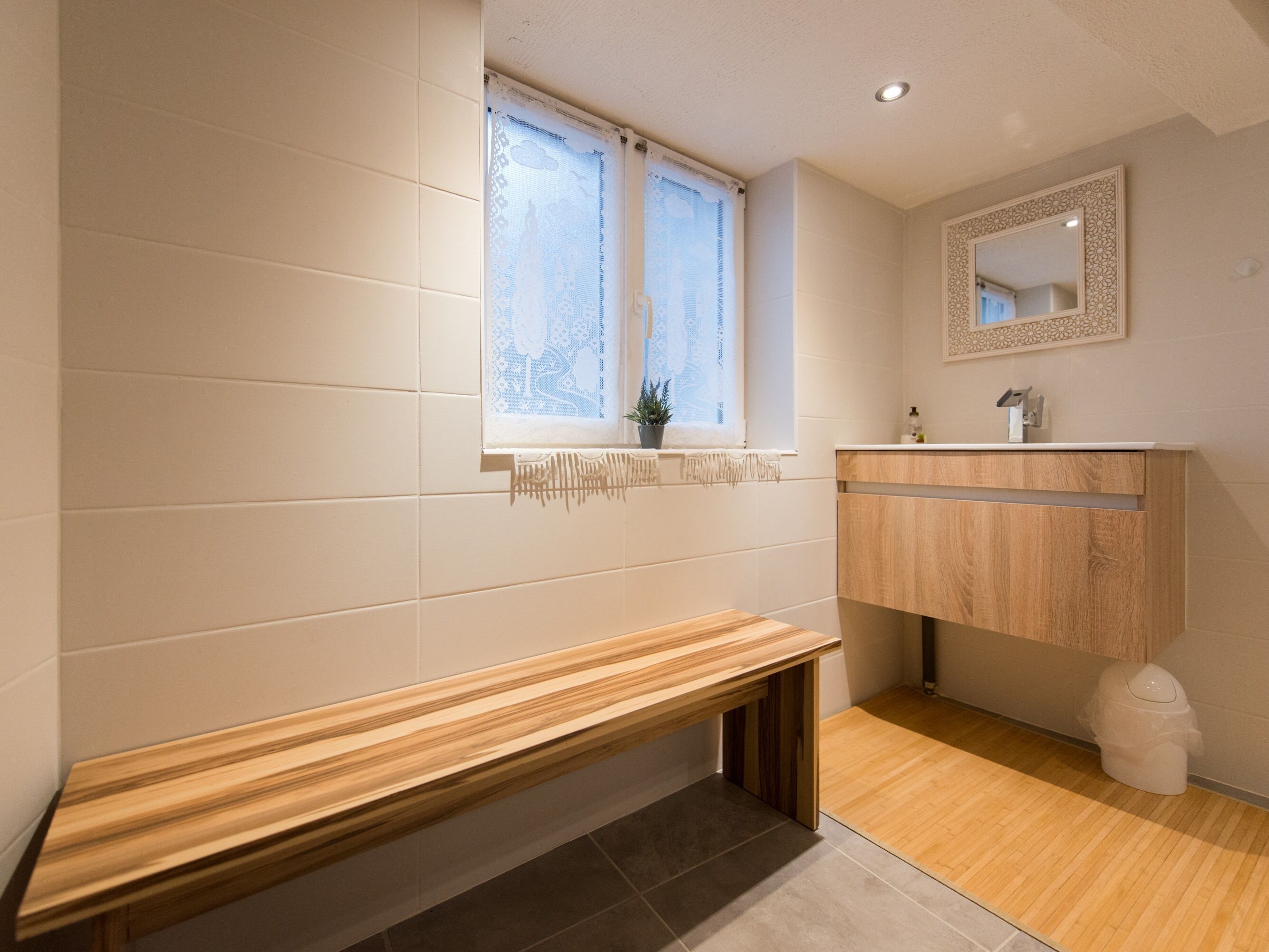 Une salle de bain avec un banc en bois, une fenêtre ornée de rideaux en dentelle, un miroir et un lavabo avec un meuble en bois.