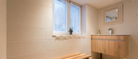 Une salle de bain avec un banc en bois, une fenêtre ornée de rideaux en dentelle, un miroir et un lavabo avec un meuble en bois.