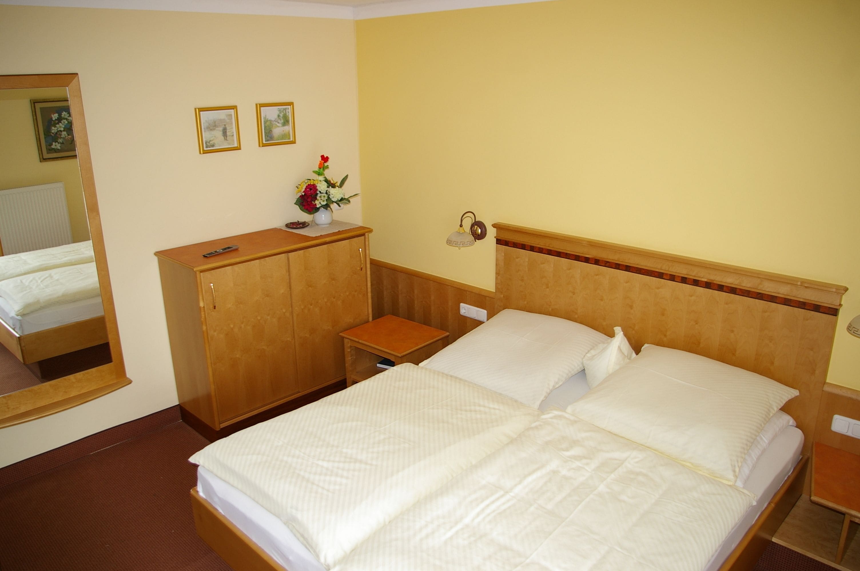 A hotel room with a double bed, wooden headboard, a small wooden nightstand, a mirror, and a wall-mounted lamp.
