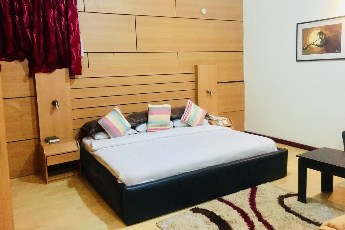 Une chambre à coucher avec une tête de lit en bois, un lit avec plusieurs oreillers, une table de chevet, une chaise et un tableau encadré au mur.