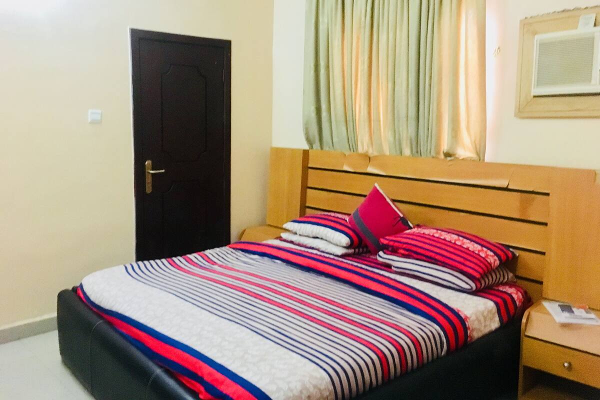 Une chambre à coucher avec un lit, une tête de lit en bois, une table de chevet, une porte et une fenêtre avec des rideaux.