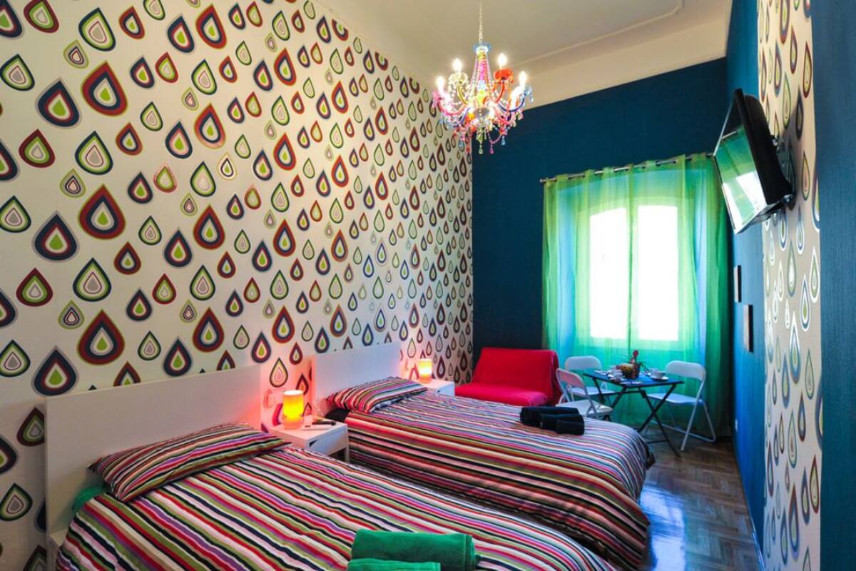 A hotel room with two beds, a chandelier, a red chair, and a TV on the wall.