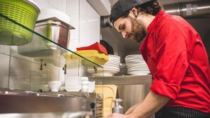Un chef, vêtu d’une chemise rouge et d’un chapeau noir, prépare des plats dans une cuisine équipée d’appareils en acier inoxydable et dont les murs sont recouverts de carreaux blancs.