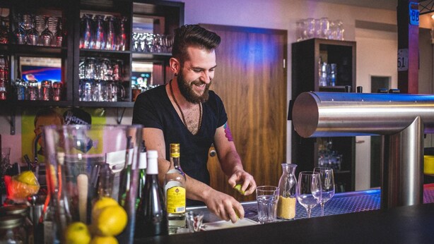 Un barman prépare des boissons dans un bar où l’on peut voir des étagères remplies de verres et de bouteilles.
