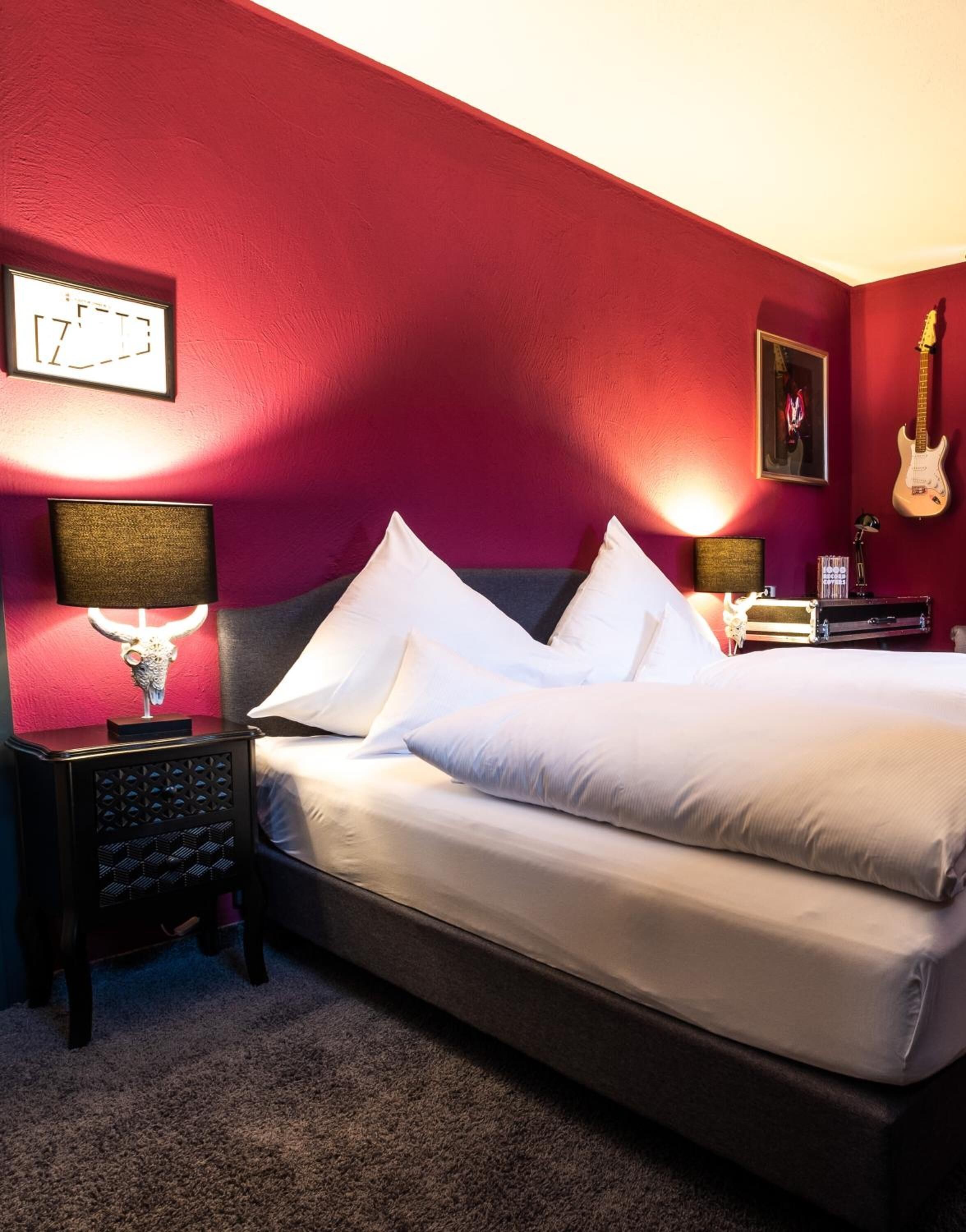 Een hotelkamer met een groot bed, een nachtkastje met een lamp, een gitaar en een ingelijst schilderij aan de muur.