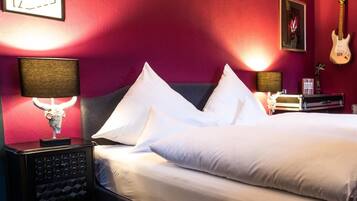 Een hotelkamer met een groot bed, een nachtkastje met een lamp, een gitaar en een ingelijst schilderij aan de muur.