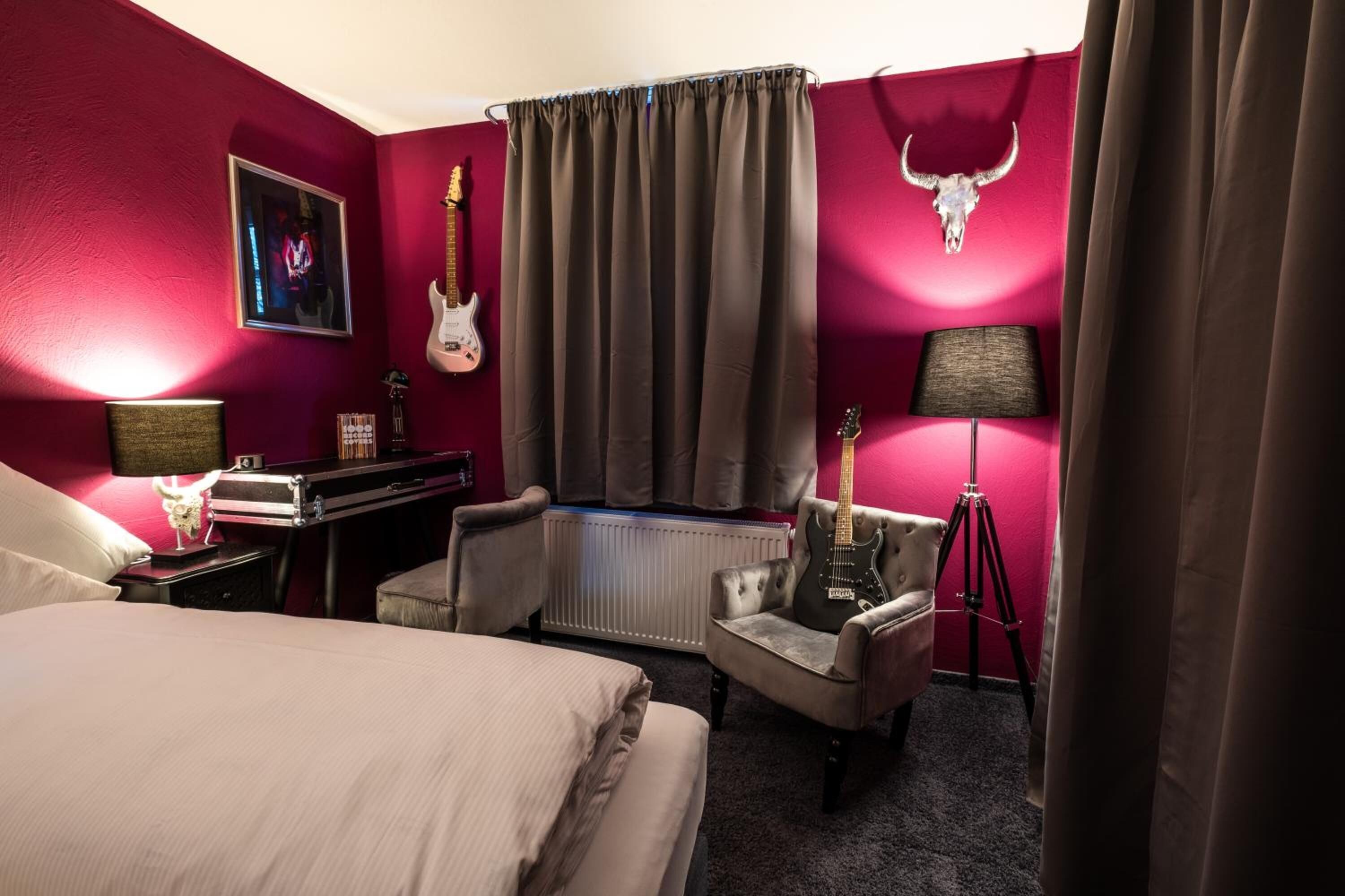 A hotel room with a large bed, a desk with a lamp, two chairs, a guitar, a radiator, and a bull skull decoration on the wall.