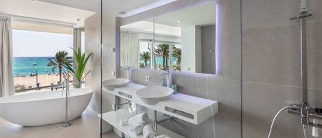 A modern bathroom with a large bathtub, two sinks, and a shower. It features a large mirror and a view of the beach.