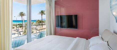 Ein Hotelzimmer mit einem großen Bett, einem an der Wand befestigten Fernseher und einem Balkon mit Blick auf den Strand und Palmen.