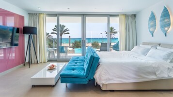Habitación de hotel moderna con una cama grande, un área de estar con vista a la playa, un televisor de pantalla plana y una mesa de comedor.