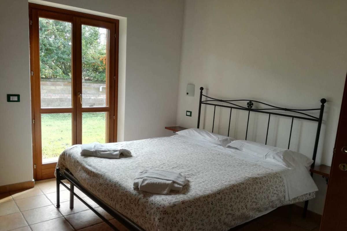 Een enkel bed met een bloemenbedekking en twee kussens, een houten deur met ruitpanelen, en een klein nachtkastje.
