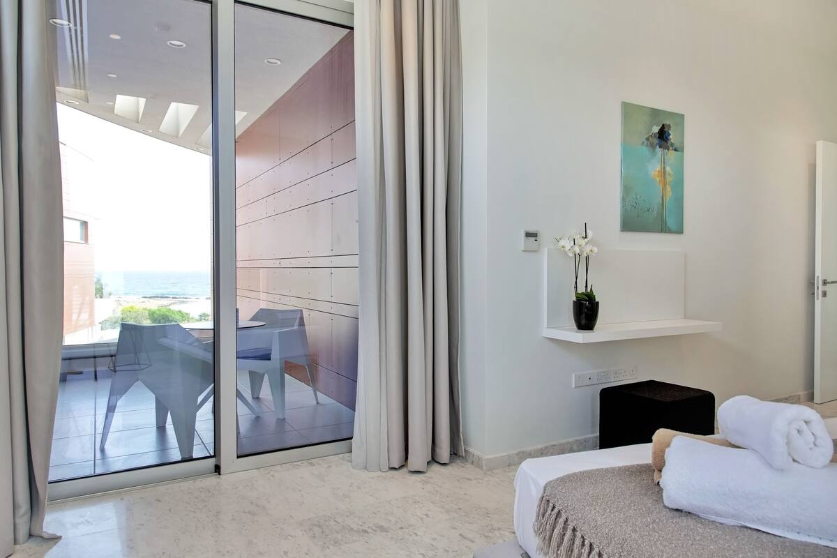 Une chambre d’hôtel moderne avec un balcon, un lit avec du linge de lit blanc, un tableau au mur et une petite plante en pot.