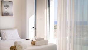 A neatly made bed with rolled towels, a bedside table with a lamp, and a large window with sheer curtains. - Pearl Beachfront Villa (Ayia Napa)