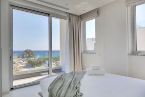 A hotel room with a large sliding glass door, a bed with white linens, a grey knitted throw, and a view of the ocean and a distant island. - Pearl Beachfront Villa (Ayia Napa)