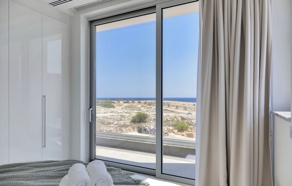 A bedroom with a large sliding glass door leading to a balcony with a view of rocky terrain and the sea.