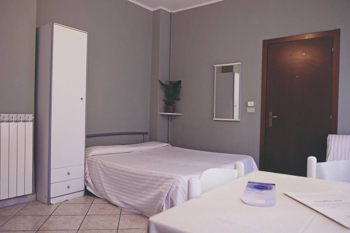 Una habitación pequeña y limpia con cama, armario, una mesita con silla, una planta en maceta y una puerta.