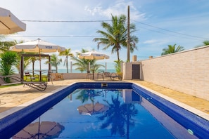 A swimming pool with lounge chairs and umbrellas, palm trees, and a view of the ocean.