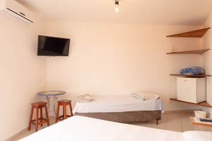 Un dormitorio pequeño y ordenado, con cama, televisor montado en la pared, una mesita con dos taburetes y un refrigerador.