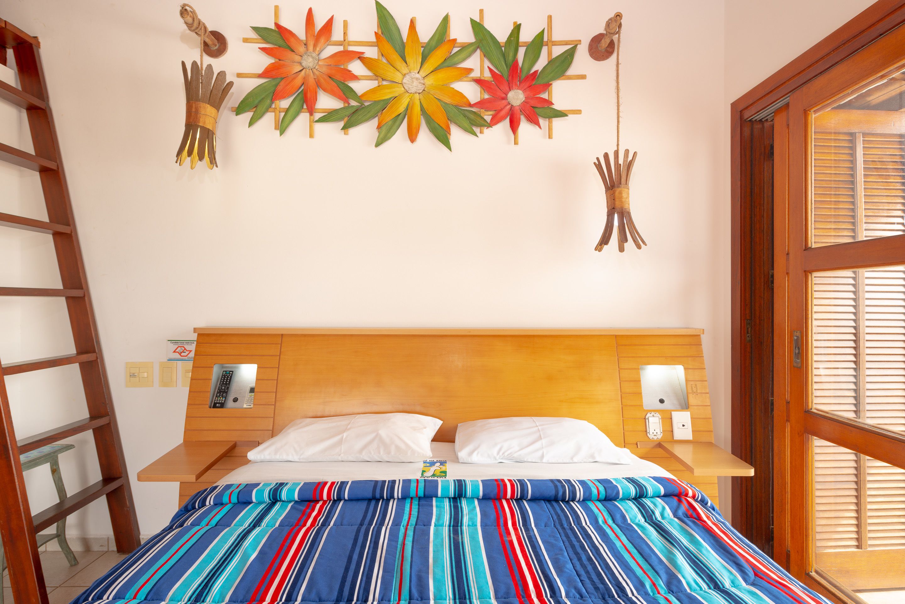 A bed with a striped bedspread, a wooden headboard, two white pillows, a bedside table with a phone and a lamp, and a wall decoration above the bed.