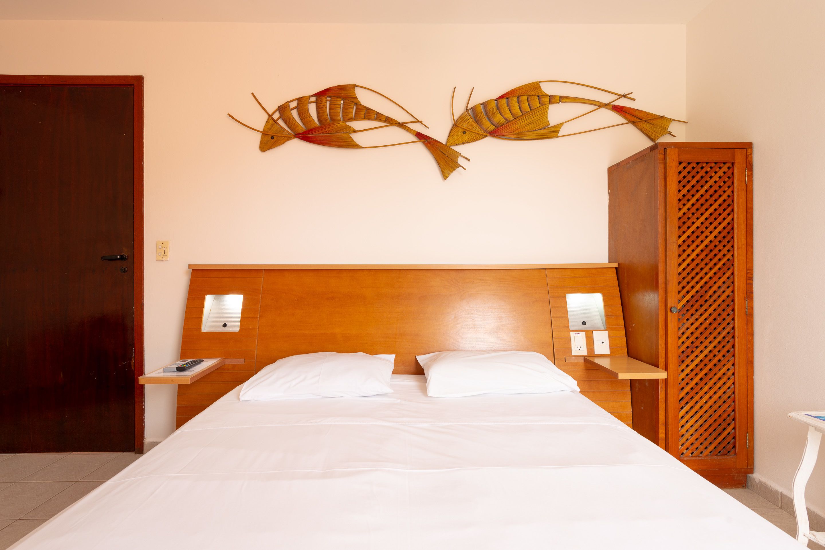 A hotel room with a wooden headboard, two white pillows, a wooden nightstand with a phone, and a decorative fish wall art piece.