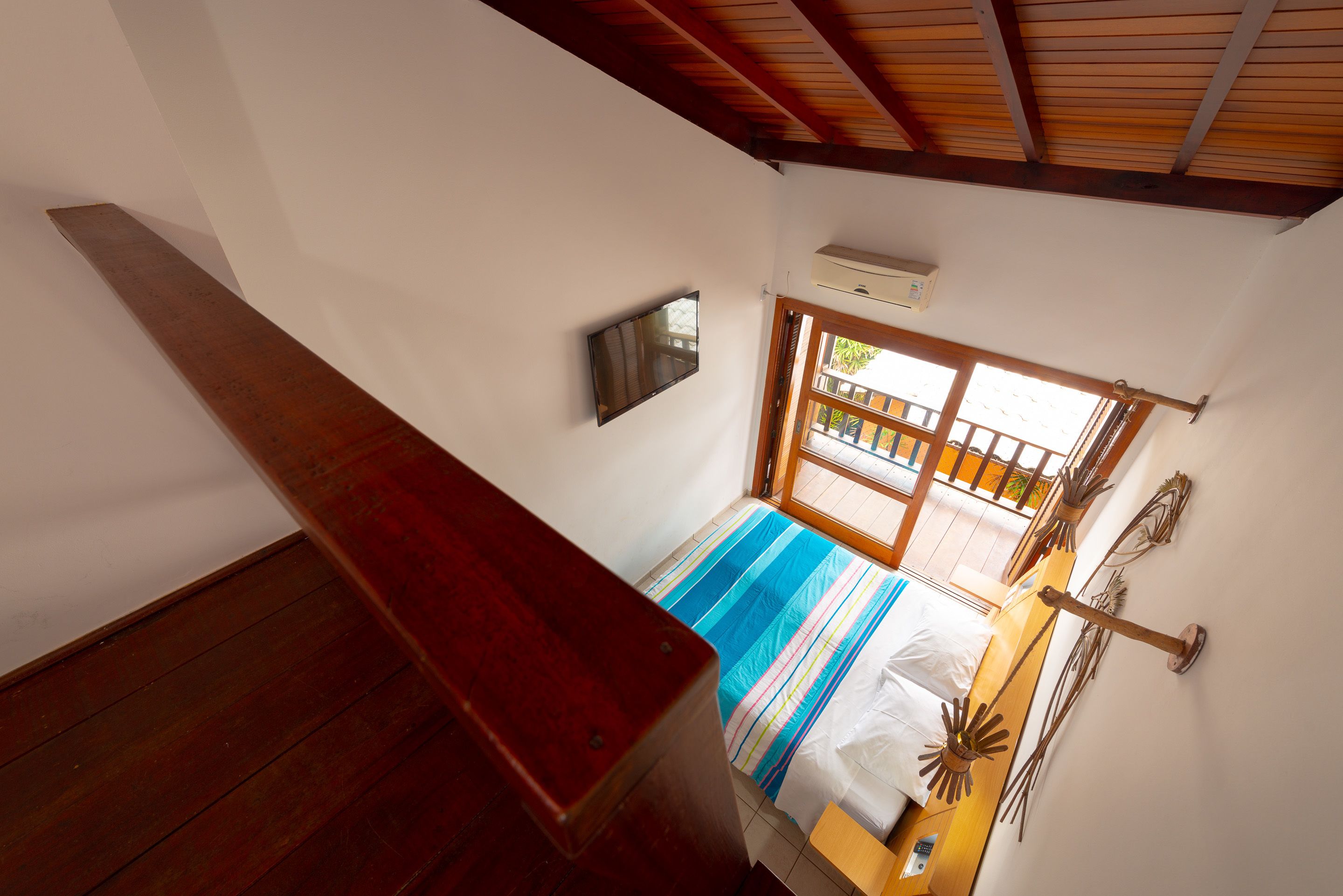 Quarto com teto de madeira, cama com colcha listrada, televisão fixada na parede e janela com vista para o exterior.