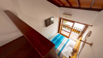 Quarto com teto de madeira, cama com colcha listrada, televisão fixada na parede e janela com vista para o exterior.