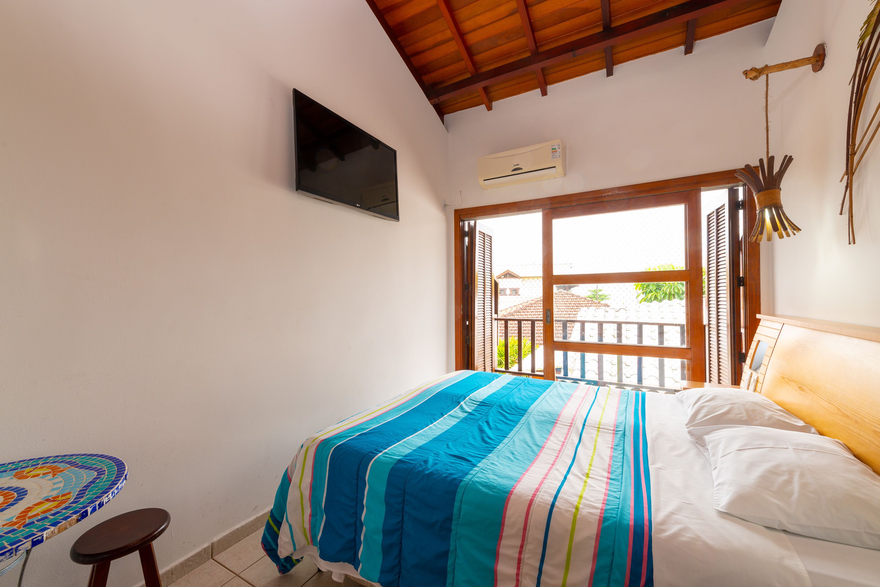 Quarto com cama, televisão, mesinha e janela com vista para um edifício e vegetação.