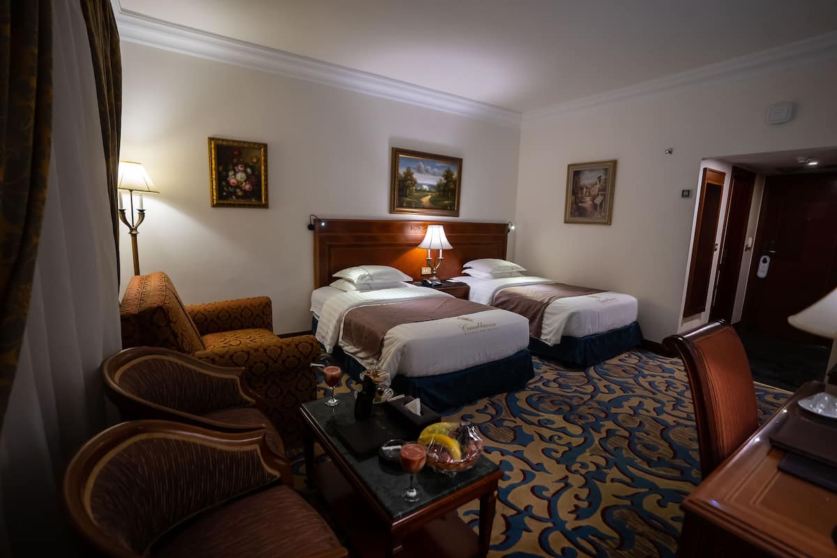 Een hotelkamer met twee bedden, een bureau met lamp, een stoel en een eettafel met een schaal fruit en glazen.