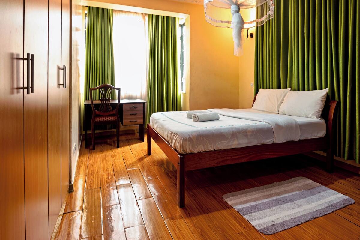 A bedroom with a wooden floor, a bed with white linens, a desk with a chair, green curtains, and a hanging light fixture.