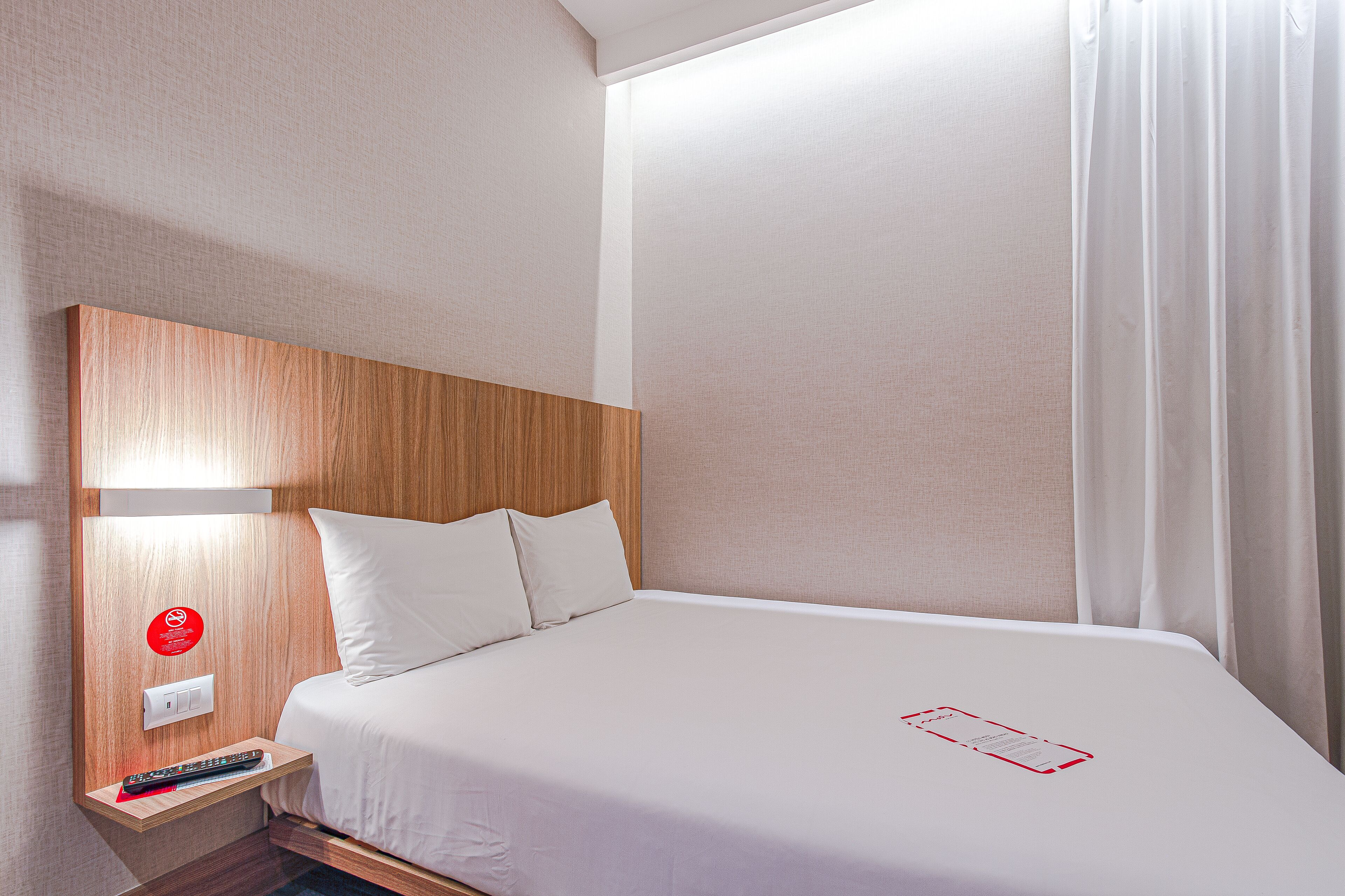 Quarto de hotel com cabeceira de madeira, cama branca, mesa de cabeceira com controle remoto e um letreiro vermelho na parede.