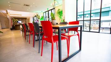 Uma área de refeições moderna com cadeiras vermelhas e uma mesa comprida, janelas grandes e plantas dentro de casa.