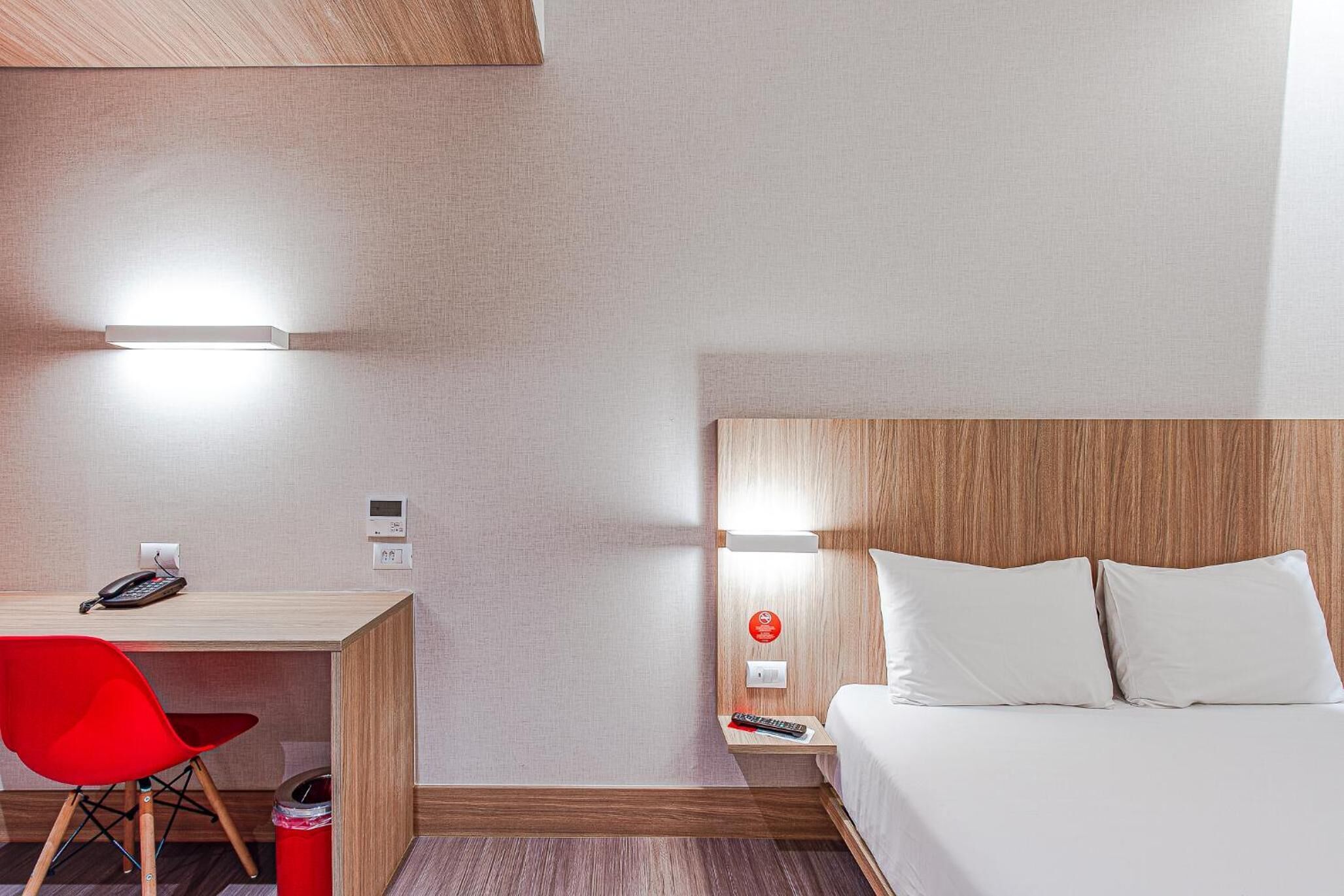 A hotel room with a wooden desk, a red chair, a bed with white linens, and a nightstand with a remote control.