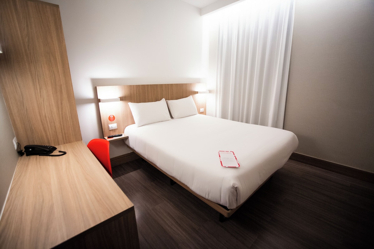 Een hotelkamer met een bed, een bureau, een stoel, een nachtkastje, een lamp en een raam met gordijnen.