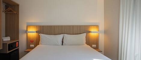 A hotel room with a large bed, two bedside lamps, a wooden headboard, a wardrobe, and a small table with a remote control.