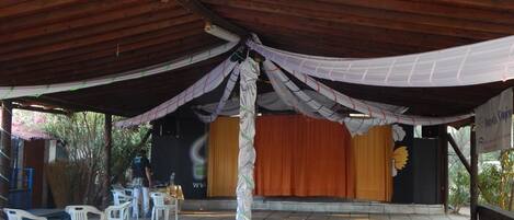 An outdoor covered area with a stage, white plastic chairs, and a wooden ceiling.