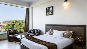 A modern hotel room with a large bed, a sitting area with a chair and table, a nightstand with a vase of flowers, a painting on the wall, and a view of greenery and buildings outside. - R G Hotels and Resort (Malur)
