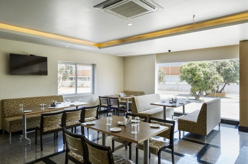 A hotel dining area with tables, chairs, and a sofa. There is a TV mounted on the wall and large windows providing natural light.