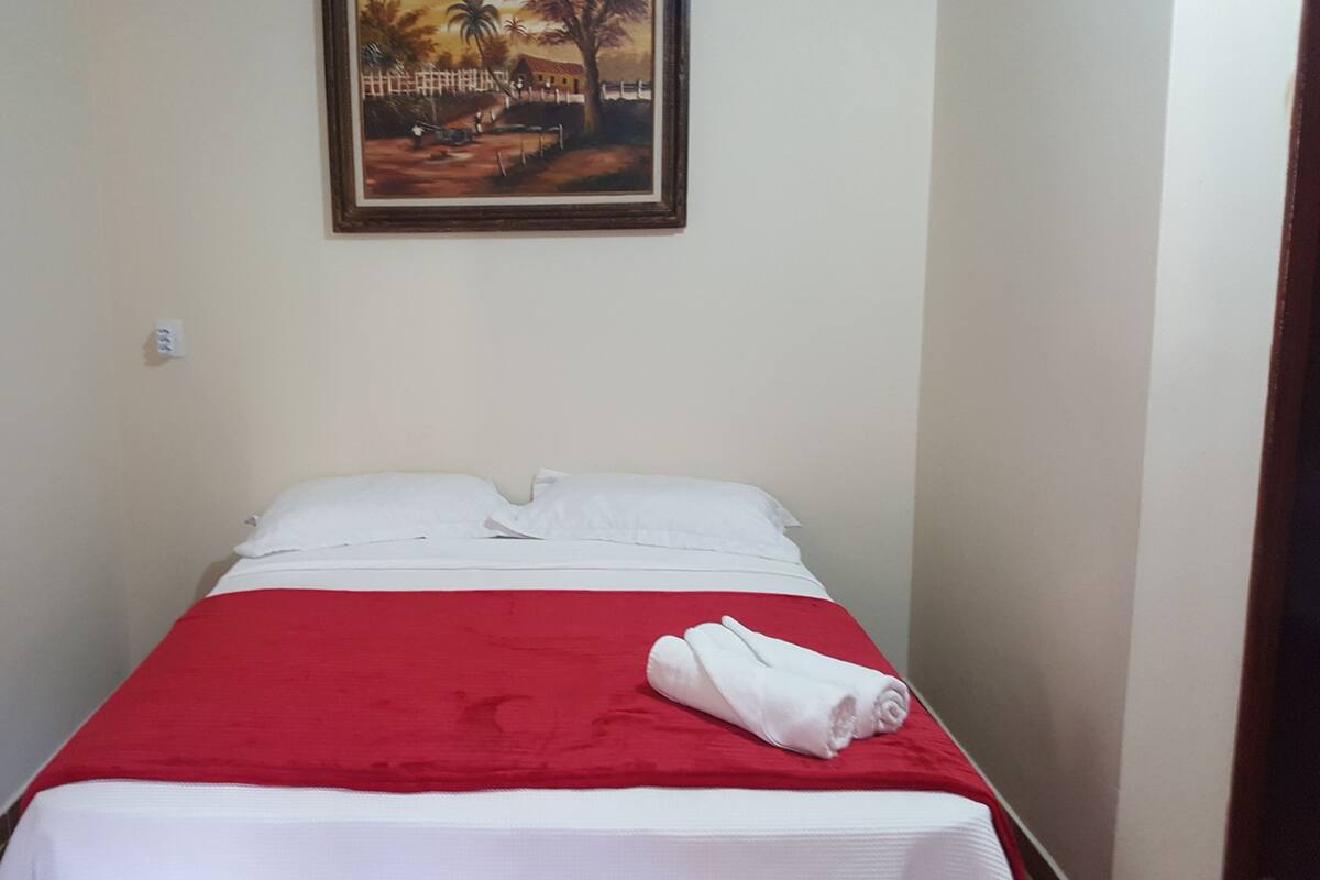 A single bed with a red bedspread and white pillows, a rolled white towel, and a painting of a rural scene on the wall.