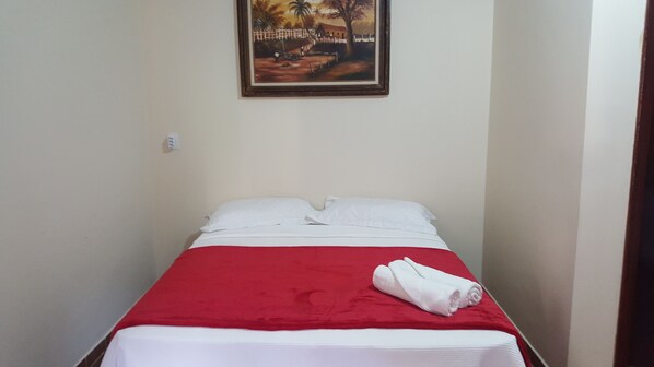 A single bed with a red bedspread and white pillows, a rolled white towel, and a painting of a rural scene on the wall.