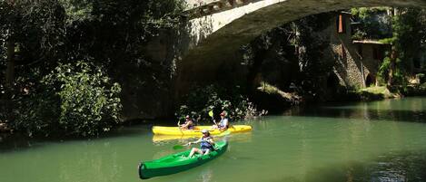 A river with kayakers under a stone bridge, surrounded by greenery.