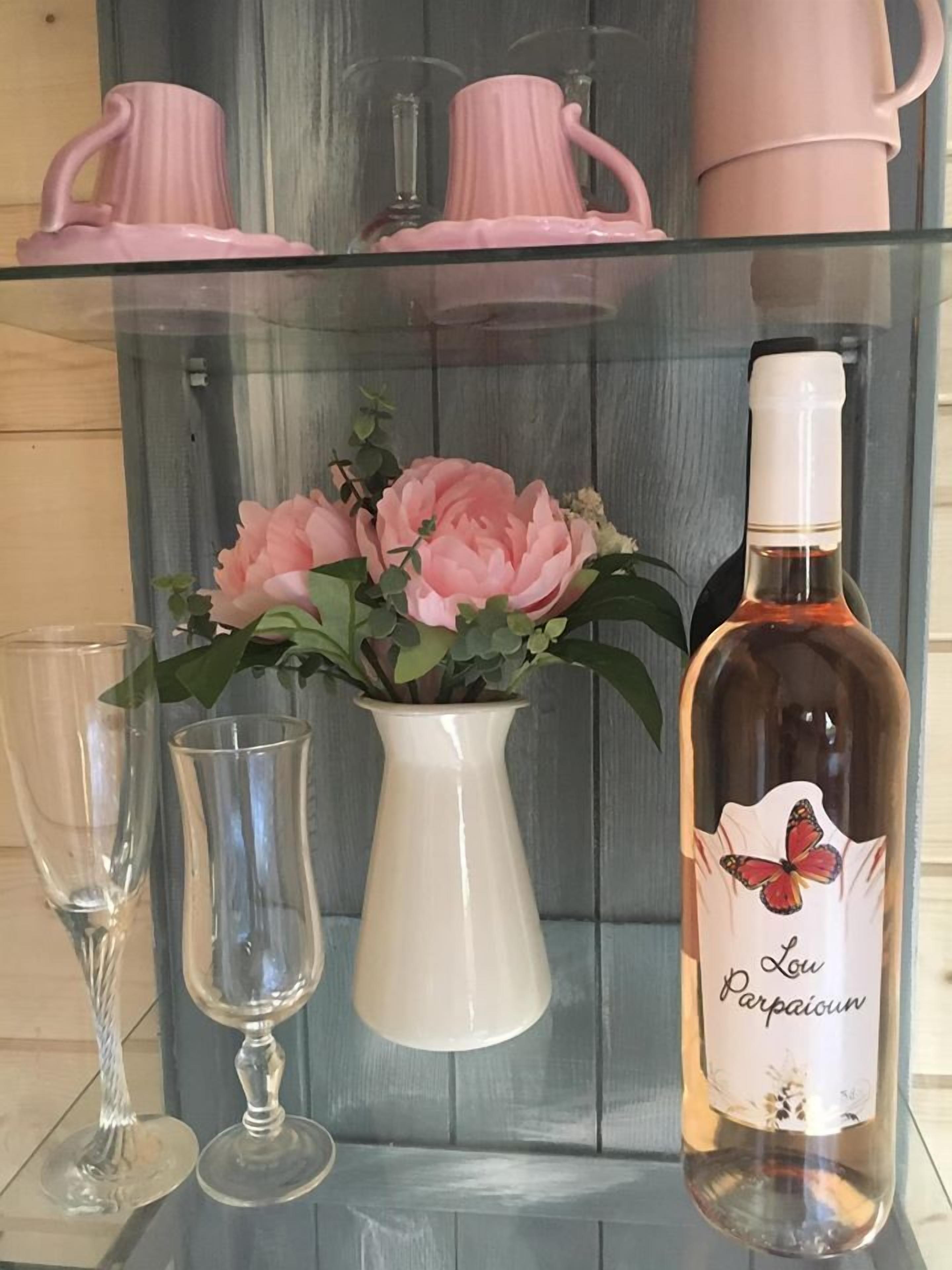 Ein gläsernes Regal mit rosafarbenen Kerbecher, einer weißen Vase mit rosa Blumen und einer Weinflasche.